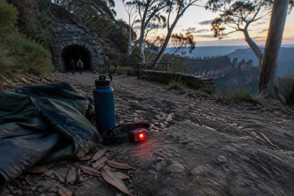 A headlamp with a red light mode lying on a rocky trail beside a water bottle and warm jacket.