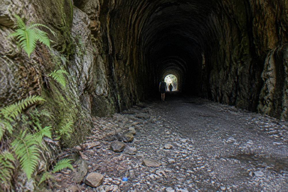 The interior of a long dark abandoned railway tunnel with uneven rocky floor and a distant pinpoint of daylight