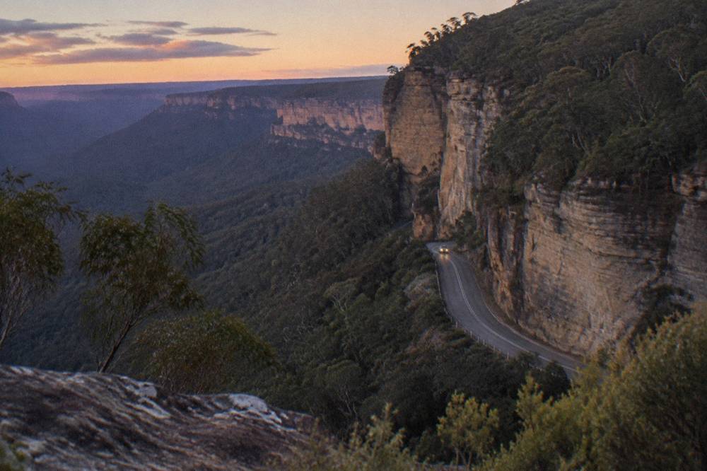 A winding mountain road at dusk with sandstone cliffs above the tree line
