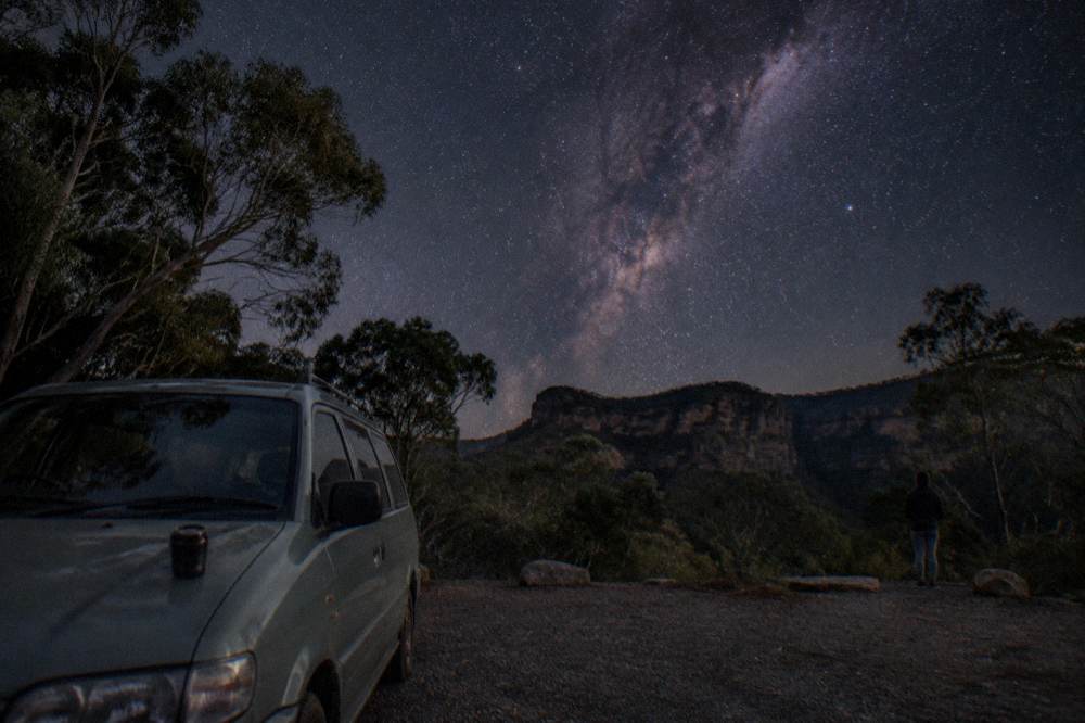 A small van parked on a dark forest road with headlights off and stars above