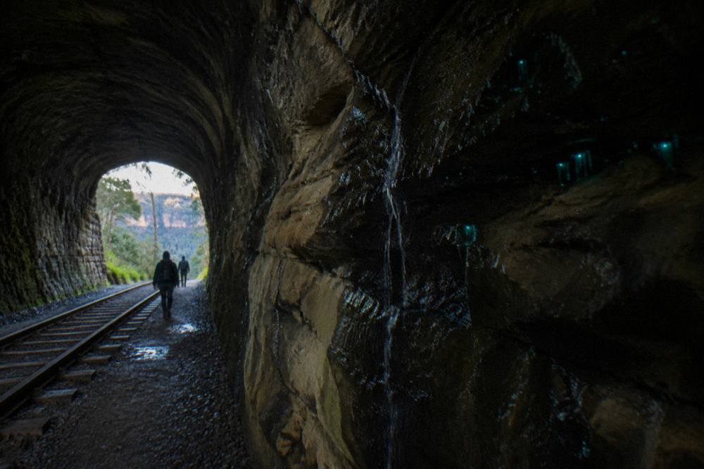 Interior of a long dark railway tunnel with a curved stone lining and faint daylight at the far end