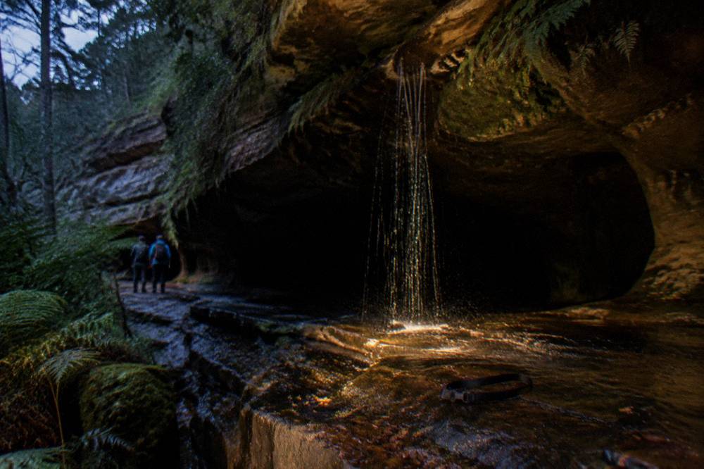A waterfall trickling over a dark cave mouth surrounded by moss and sandstone