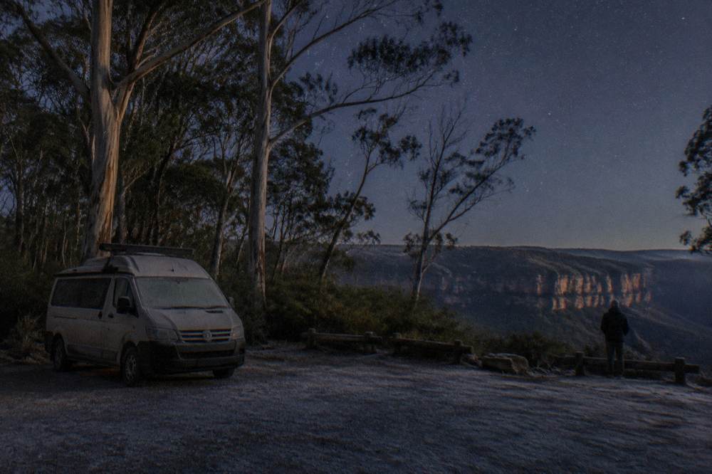 A small van parked on a dark rural road edge near tall eucalyptus trees at night