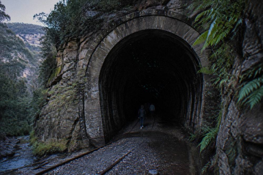 Dark abandoned railway tunnel with a curved stone lining and faint blue-green lights along the ceiling