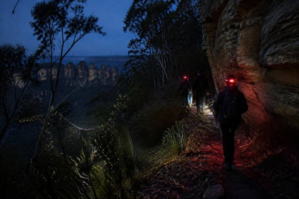 Headlamps and red lights on a dark bush track beside a rock face at night