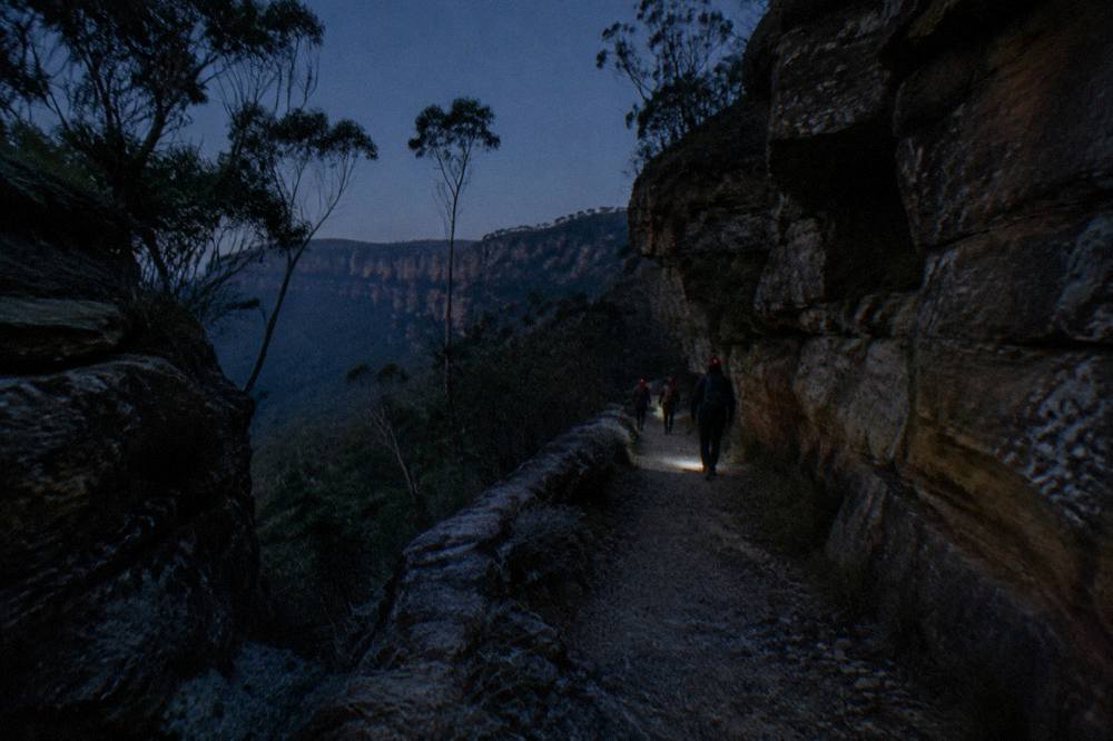 A small group of hikers with red headlamps walking a dark bush track between sandstone walls