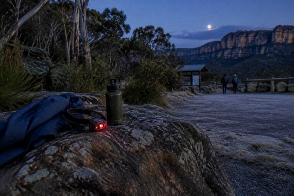 A headlamp with a red glow resting on a rock beside a water bottle and jacket