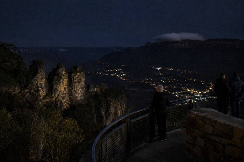 A night sky over dark sandstone cliffs with the faint lights of a distant town in the valley below