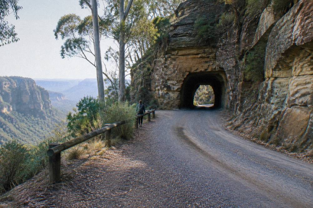 A narrow gravel road curving between sandstone cliff walls with a single-lane tunnel opening visible ahead