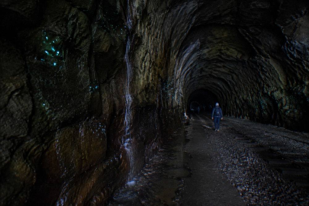 A dark railway tunnel interior with rough stone walls curving into the distance, faint green light visible deep inside