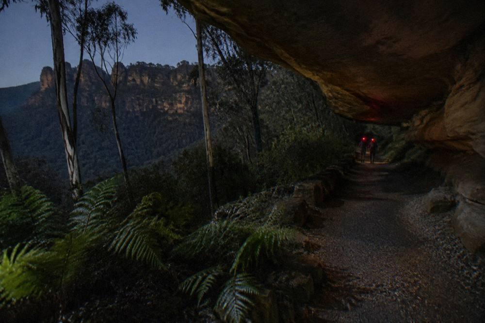 Headlamps glowing red along a dark bush track beside a rock overhang at night