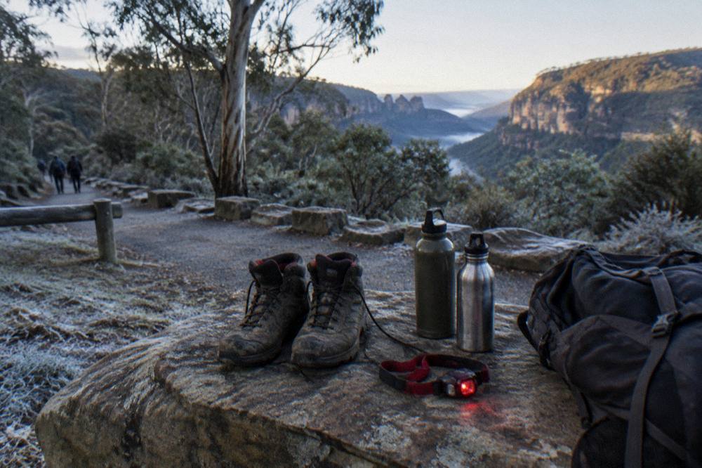 Walking gear laid out on a timber bench at a trailhead: boots, water bottles, a red-light headlamp