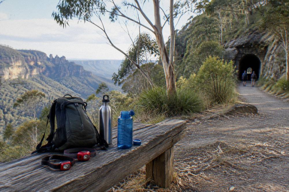 A daypack, water bottles and headlamps arranged on a wooden bench beside a forest walking track