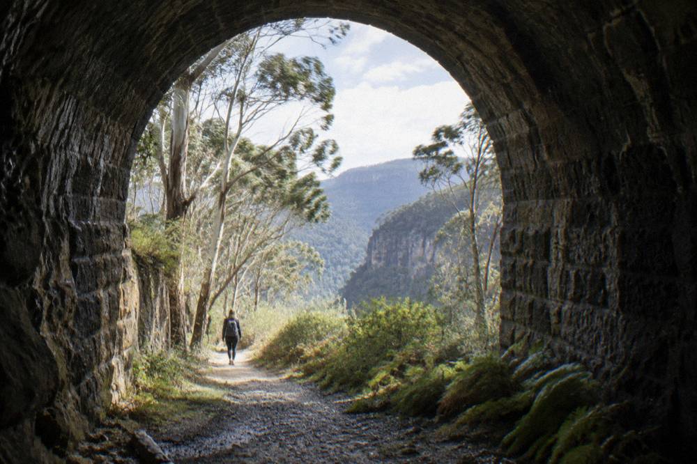 A curved disused railway tunnel of rough stone opening onto a bush walking track
