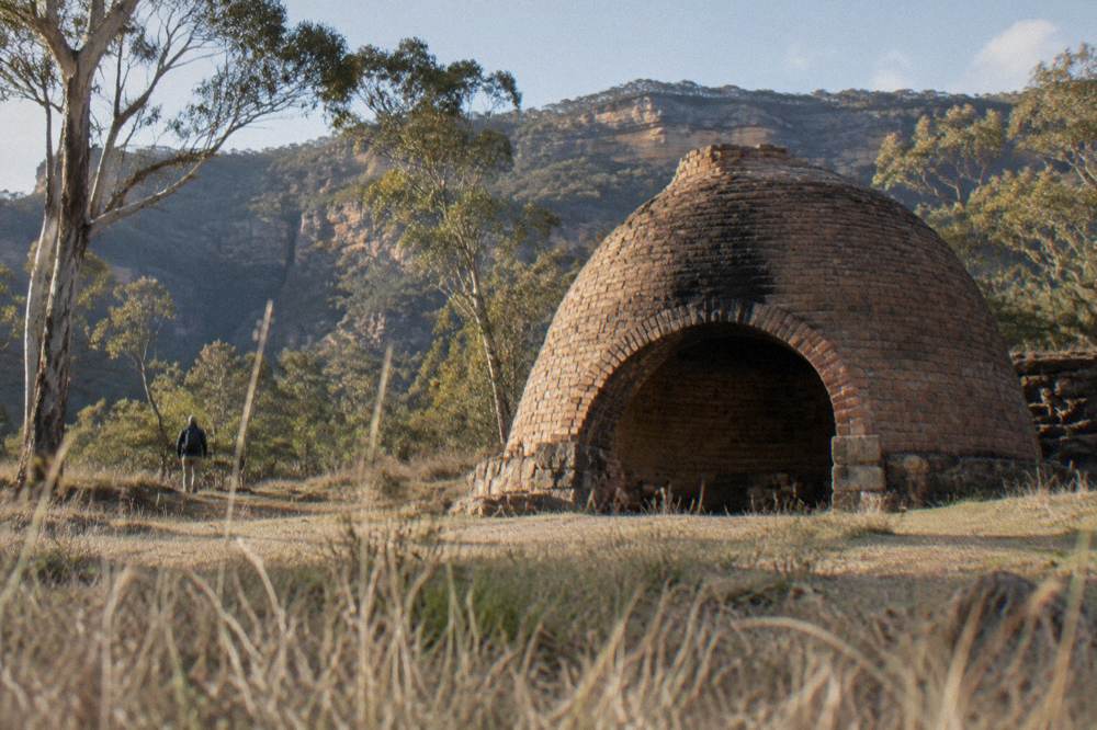 A domed brick kiln standing open in a grassy clearing with forested ridges behind