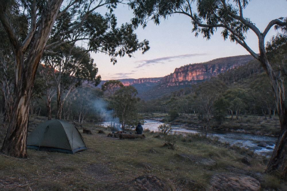 A quiet riverside campsite among trees with sandstone cliffs in the distance