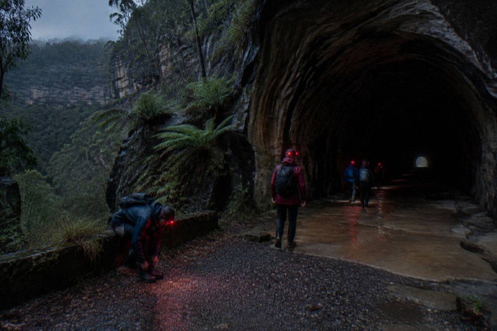 A small group of walkers with red headlamps entering a dark railway tunnel cut through sandstone