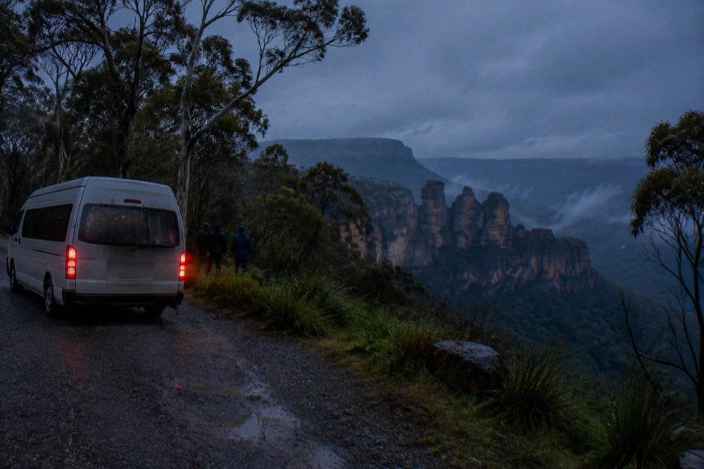 A small van parked at night on a bush road with headlamps glowing near the treeline