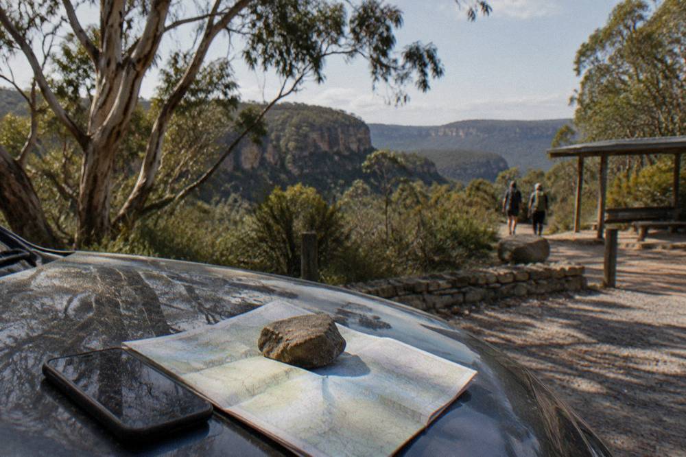 A smartphone showing an offline map beside a paper topographic map on a car bonnet in forest