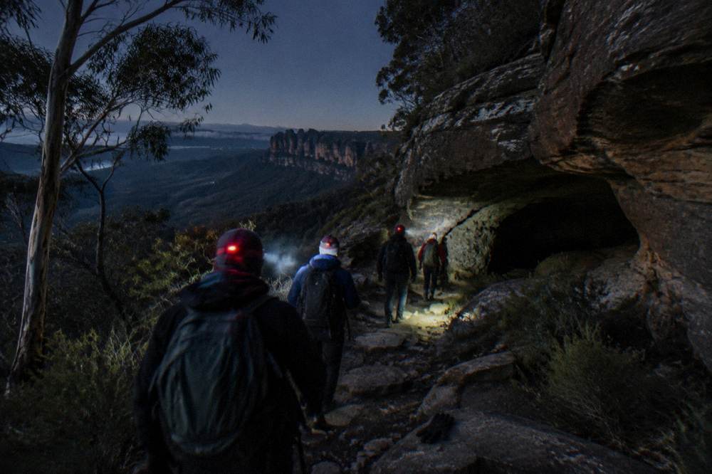 A small group of hikers with red headlamps walking a dark bush track toward a rocky cave entrance at night